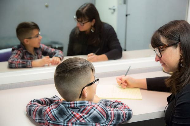 mirror image of a young boy with glasses wearing red and black flannel sitting at a desk with an adult female with glasses taking notes on a large yellow notepad