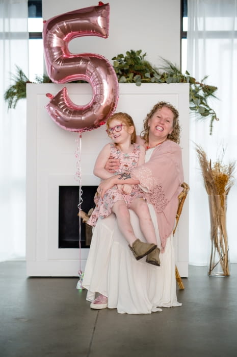 white room, woman holding child on her lap, smiling at camera, with a pink number 5 balloon to the left of them