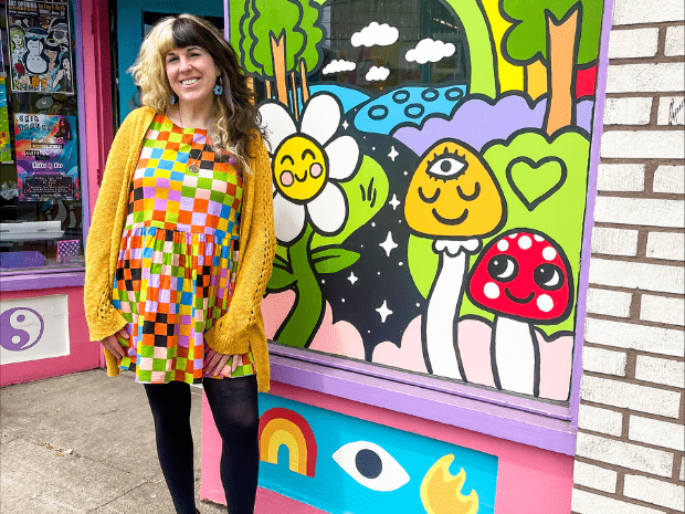 Person with split dye hair, blonde on left and black on right, wearing a colorful geometric dress with black leggings and a yellow cardigan, posing outside of a business window with colorful paintings of flowers and mushrooms
