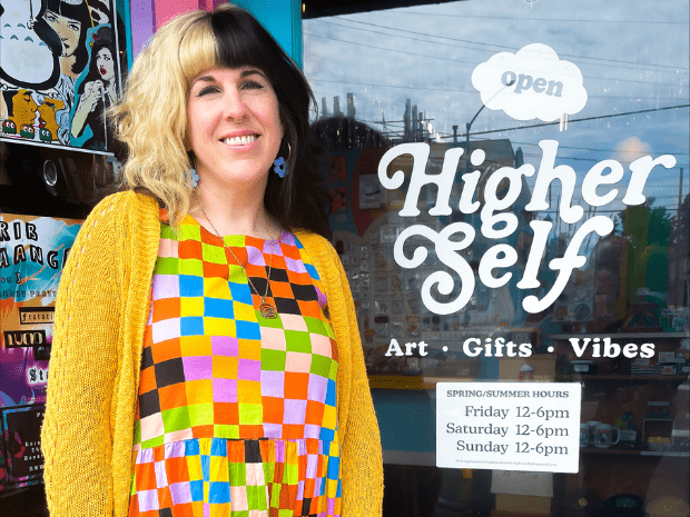 Person with split dye hair, blonde on left and black on right, wearing a colorful geometric dress with black leggings and a yellow cardigan, posing outside of a glass business door with Higher Self inscribed on the door, a white open sign, and a white sign featuring business hours