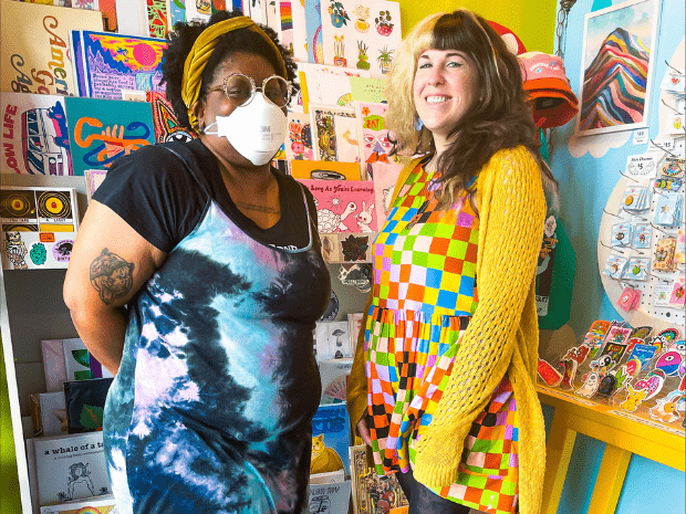 Person on the left is wearing a white medical face mask with white round glasses, and a black shirt under a blue and teal tie-dye dress. Person on the right with split dye hair, blonde on left and black on right, wearing a colorful geometric dress with black leggings and a yellow cardigan