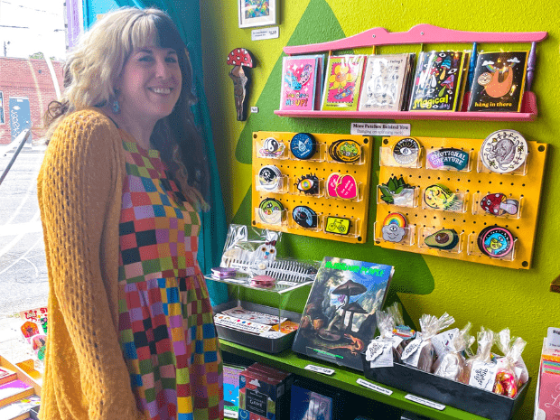 Person with split dye hair, blonde on left and black on right, wearing a colorful geometric dress with black leggings and a yellow cardigan, posing next to some stickers in their store