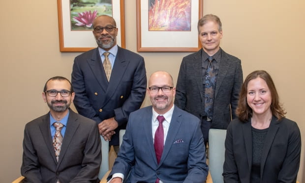 five individuals in business professional attire posing for photo, two men in background, two men in foreground, one woman in foreground to the right