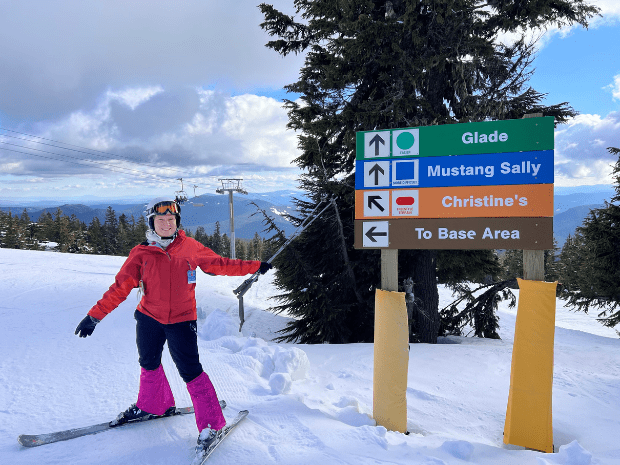 Christine Barlow Reed wearing a red ski jacket and black and pink snow pants, with skis, posing on a snowy mountain, pointing a ski pole at a colorful sign that reads "Christine's"