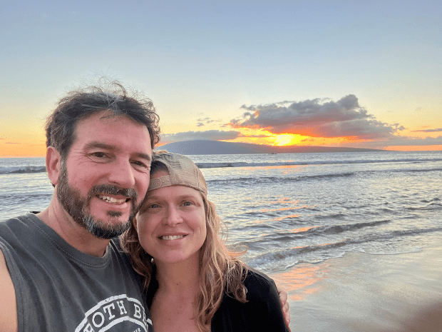 Christine Barlow Reed and husband, smiling, posing in front of a sunset on a beach
