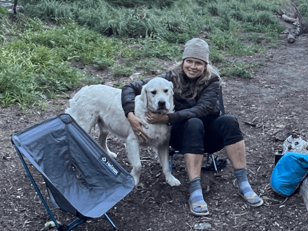 Christine Barlow Reed wearing a knit cap, sitting in camp chair in a dirt clearing outdoors, hugging a big white dog