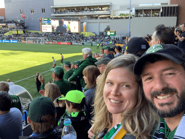 Christine Barlow Reed and husband smiling posing for camera while in the audience of a soccer game