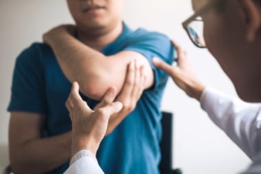 individual wearing blue shirt, holding up their left arm with their right hand. a doctor with glasses wearing a white long sleeve coat is examining the individual's elbow.