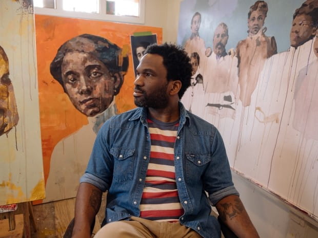 Jeremy Davis looking to the left, sitting in a studio surrounded by colorful paintings