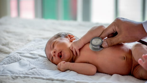 newborn laying on white sheet, with a doctor gently pressing a stethoscope on the newborn's abdomen