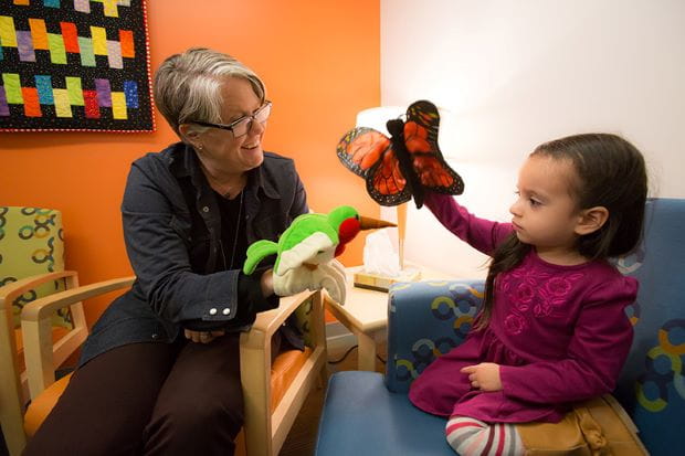 young girl wearing a purple dress playing with a red and black butterfly toy sitting next to a seated elderly adult woman with glasses with a green hummingbird plush toy